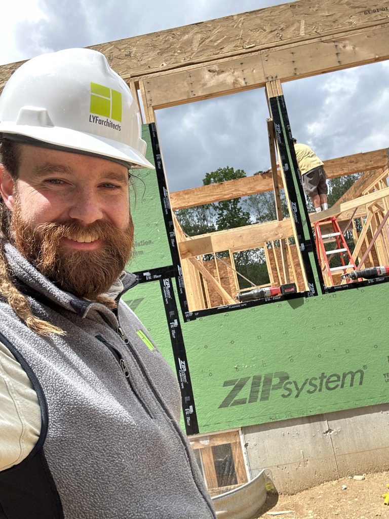 Photo of Galen Anderson wearing a hardhat in front if a building site. The building is a wood framed home with green Zip System sheathing. 